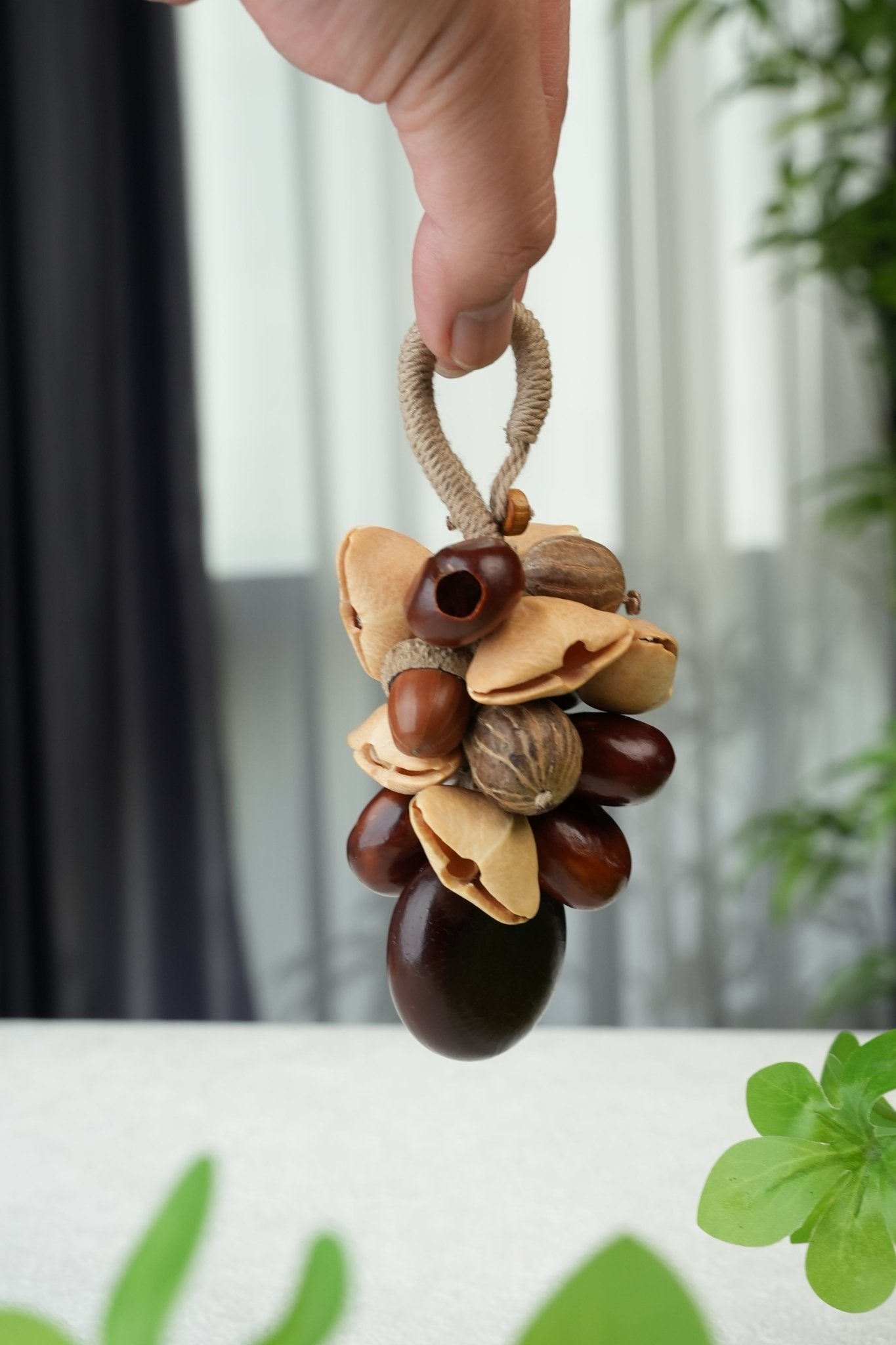 Mixed nut and seed cluster bag charm with chestnuts, pistachio shells and acorn cap on jute loop, held indoors against green plants